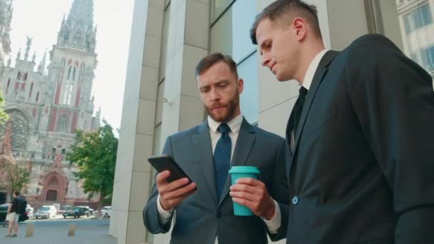 Two male employees interacting with each other in downtown business district. Confident businessmen using smartphone discussing planning computer business project brainstorm in corporate teamwork - Footage, Video