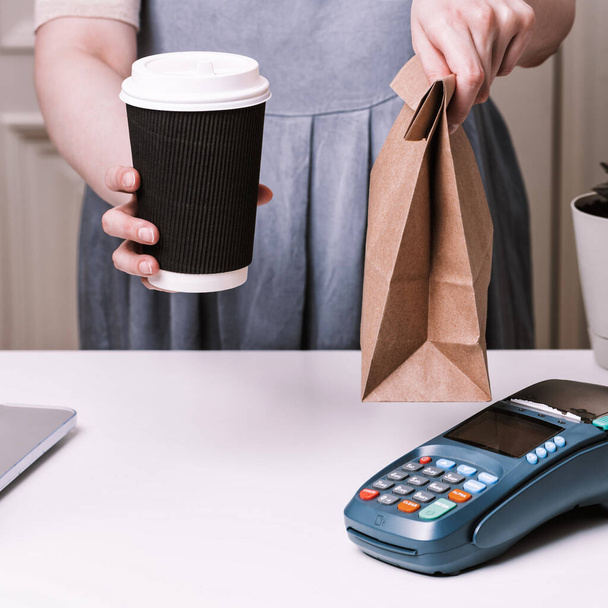 Woman holding ordering in a cafe. NFC payment terminal and take away cup of coffee with on a white table. Credit card or phone pay pos banking device, card machine. - Photo, Image