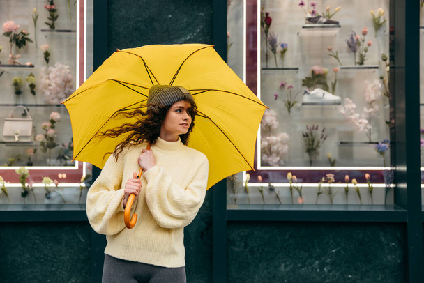 Viehättävä nuori kihara nainen käyttää keltaista sateenvarjoa megapolisin kadulla sateisena päivänä. - Valokuva, kuva