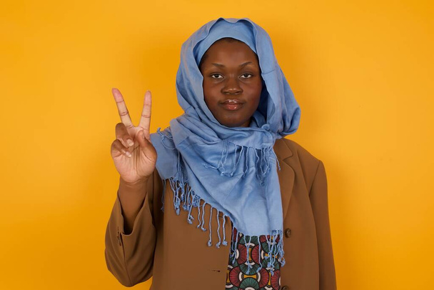 Genç Afrikalı Amerikalı Müslüman kadın gri duvarın önünde duruyor ve iki numaralı parmaklarıyla işaret ediyor kendinden emin ve mutlu gülümserken.. - Fotoğraf, Görsel