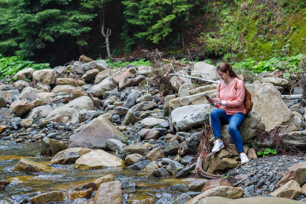 A woman works on the phone sits on a stone near a mountain river. - Φωτογραφία, εικόνα
