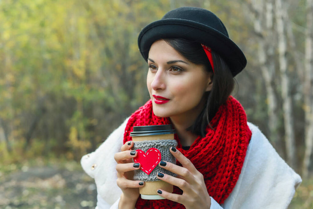 Eine junge Frau mit schwarzem Hut trinkt Kaffee aus einer gemütlichen Tasse in der Natur. Strahlendes Lächeln und gute Laune - Foto, Bild