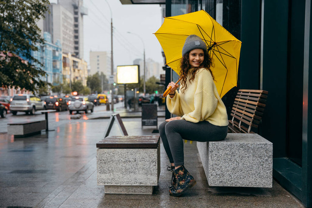 Charming young curly  woman sitting on bench and using yellow umbrella at the street of megapolis city in rainy day - Photo, Image