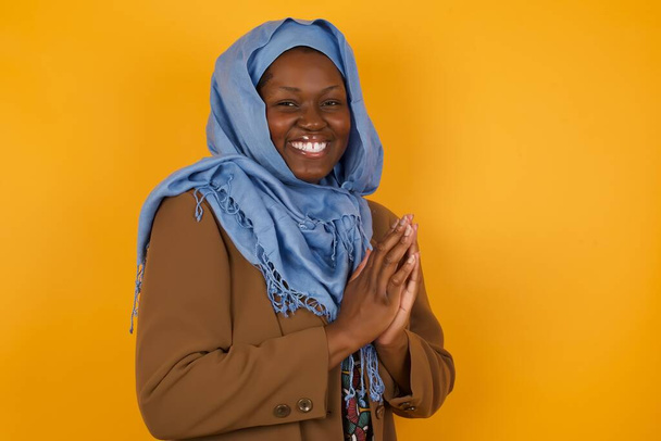Joven mujer musulmana afroamericana exitosa de pie contra la pared amarilla, sintiéndose feliz, sonriendo y aplaudiendo, diciendo felicitaciones con un aplauso. - Foto, Imagen