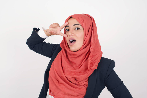 Kaunis musliminainen pukeutuu musta bleiseri ja hijab osoittaa tehdä v-merkki lähellä silmiä yllään rento vaatteita seisoo ulkona. - Valokuva, kuva
