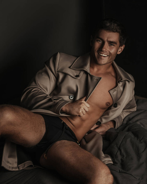 Lächelnder Mann in Mantel und Unterwäsche liegt auf dunklem Hintergrund - Foto, Bild
