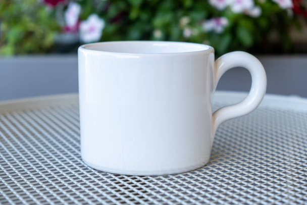 Kaffeetassen-Attrappe auf einem Gartentisch. Weiße Farbe leere Tasse mit Henkel für Heißgetränk, Blühende Blumen Hintergrund verschwimmen. Werbung, Branding-Vorlage - Foto, Bild