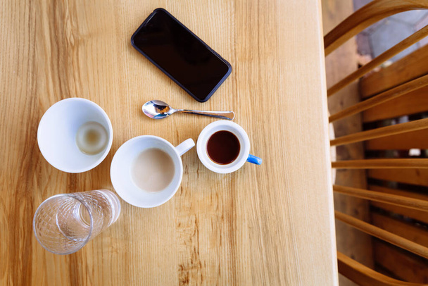 Auf einem Holztisch stehen schmutzige Kaffeetassen, Cappuccino und ein Glas Wasser. Neben den Tassen steht ein Telefon. Kaffeepause. Flache Lage - Foto, Bild