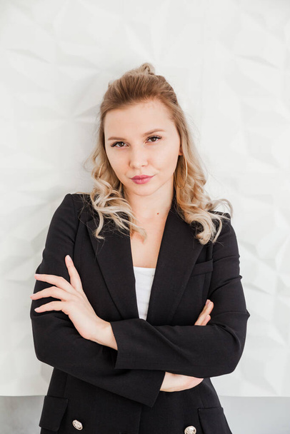 A blonde girl in a black suit stands against a white wall with crossed arms and looks into the camera. Vertycal photo - Foto, imagen