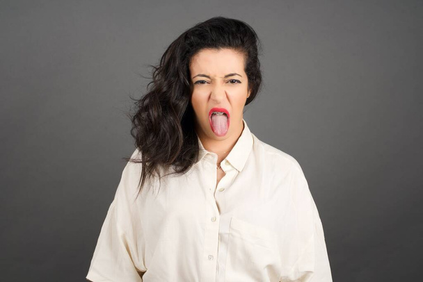 Beautiful  brunette woman dressed casually showing tongue . Standing against gray background.   - Photo, Image