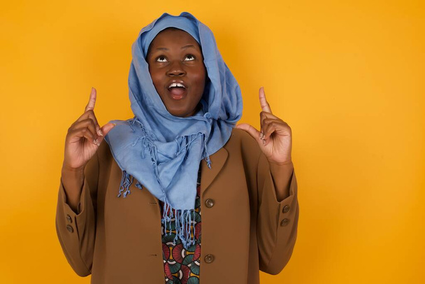 Encantadora mujer musulmana afroamericana despreocupada con expresión positiva, señala con ambos dedos índice, vestida con ropa casual, tiene una amplia sonrisa interesada, aislada sobre un fondo amarillo. Mira ahí, por favor.. - Foto, imagen