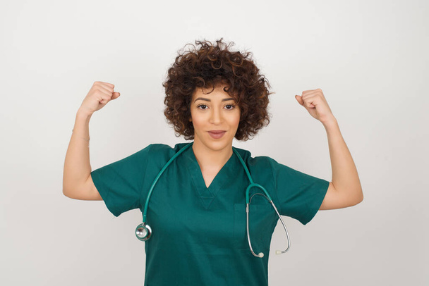 Taillierte Aufnahme der schönen arabischen Ärztin in medizinischer Uniform hebt die Arme, um zu zeigen, dass sich ihre Muskeln siegessicher fühlen, stark und unabhängig wirkt, positiv in die Kamera lächelt, vor grauem Hintergrund steht. - Foto, Bild