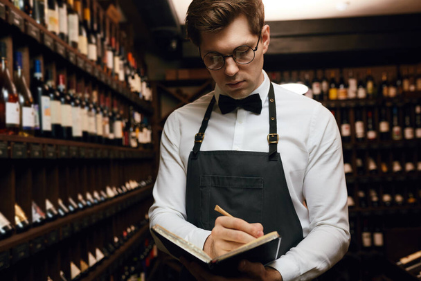 Male cavist examines the bottles with wine and making notes at his notepad - Фото, изображение