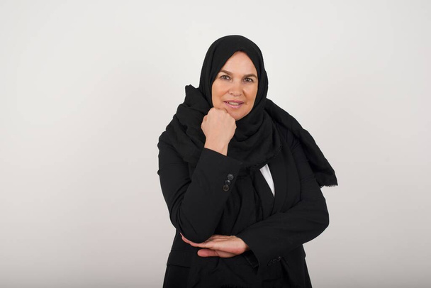 Isolated portrait of muslim woman in black hijab with hand near face and looking to the camera with doubtful and skeptical expression suspect and doubt. Isolated over gray background. - Foto, afbeelding
