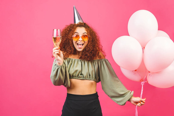 Šťastnou dovolenou! Šťastná mladá kudrnatá žena s balónky izolovanými přes růžové pozadí. Na Valentýna nebo na ženský. Držím sklenici šampaňského. - Fotografie, Obrázek