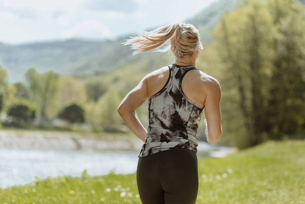 Back view photo of young blonde girl wearing sportswear running in the nature, by the river in the morning. Sunny weather. Healthy lifestyle and sport concept - Фото, изображение