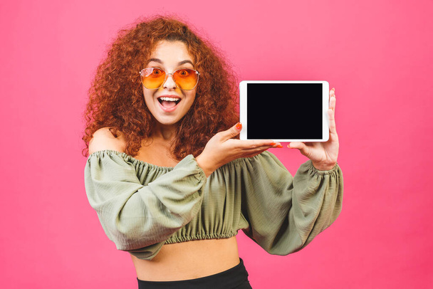 Smiling curly model with tablet computer in studio. Showing at tablet display with copy space. Isolated over pink background. - Foto, Imagem