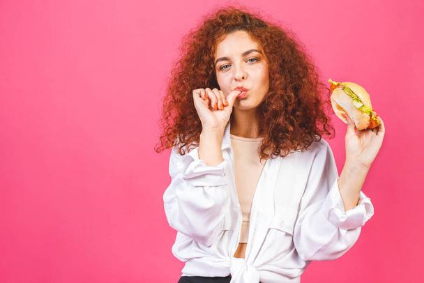 Junk Food Konzept. Attraktive junge lockige Kaukasierin, die einen großen Hamburger hält und isst, isoliert auf rosa Hintergrund.  - Foto, Bild