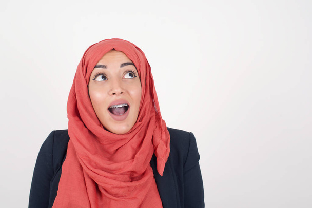 Beautiful muslim woman  wears black blazer and hijab shrugs shoulders, looking sideways, being happy and excited about new plans with friends. Isolated over gray wall. - Foto, imagen