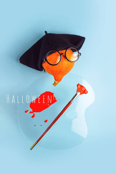 Artista de calabaza decorativa naranja en boina negra, gafas, con pincel, paleta y pintura roja sobre fondo azul. Concepto Halloween. - Foto, imagen