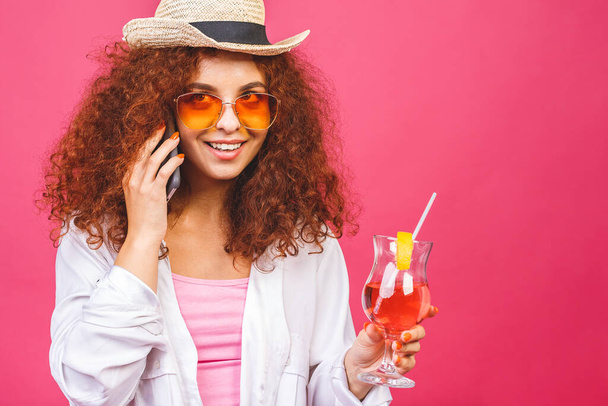 Portrait of happy woman in sunglasses using smartphone and holding cocktail isolated on pink background.  - Фото, изображение