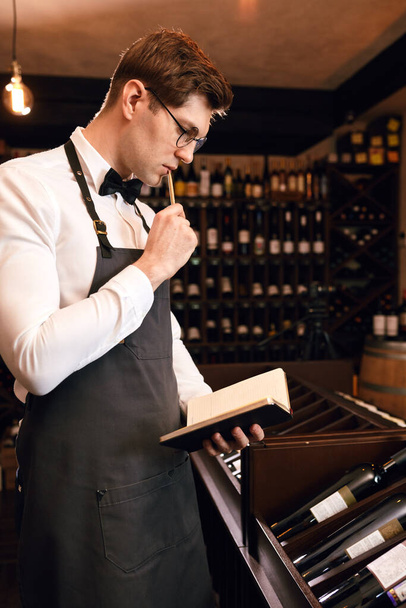 Wine steward looking at bottles of wine and writing in notebook at wine cellar - Foto, Bild