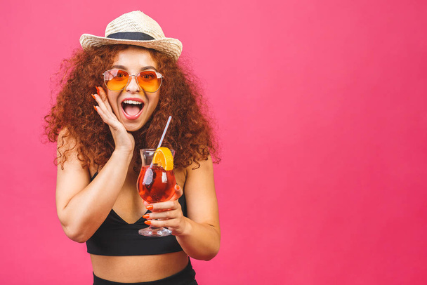 Renkli pembe arka planda izole edilmiş bir bardak kokteyl içeceği stüdyosuyla yaz kıyafetleri içinde mutlu, güzel kıvırcık kadın.. - Fotoğraf, Görsel