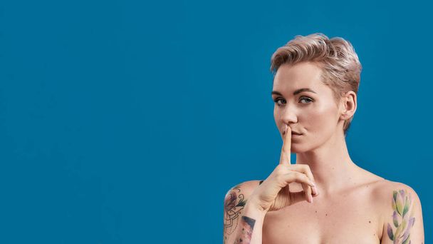 Portrait of a young attractive half naked tattooed woman with perfect skin looking at camera, holding a finger on her lips isolated over blue background - 写真・画像
