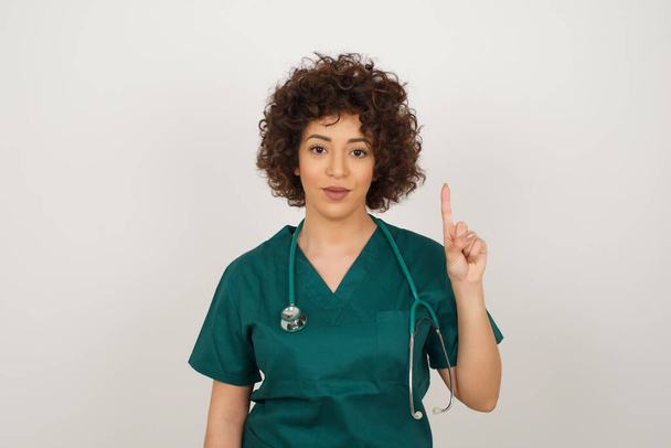  hermosa mujer médico árabe vistiendo uniforme médico, de pie contra la pared gris mostrando y señalando hacia arriba con los dedos número uno  - Foto, imagen