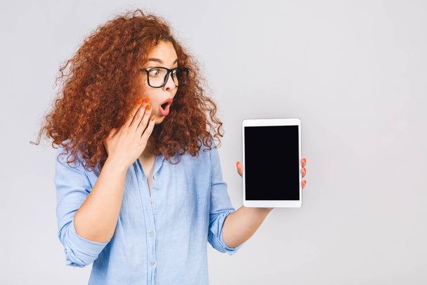 Image of cheerful amazed young curly caucasian woman showing display of tablet computer. Digital tablet screen isolated over white background. - 写真・画像