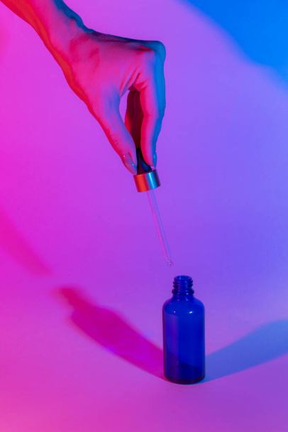 Bottle of blue on a neon trend background. Female hand holds a glass dropper. Gradient from pink to blue. Two shadows of different colors. Dropper glass Bottle Mock-Up. Cosmetics concept. - Foto, Imagen