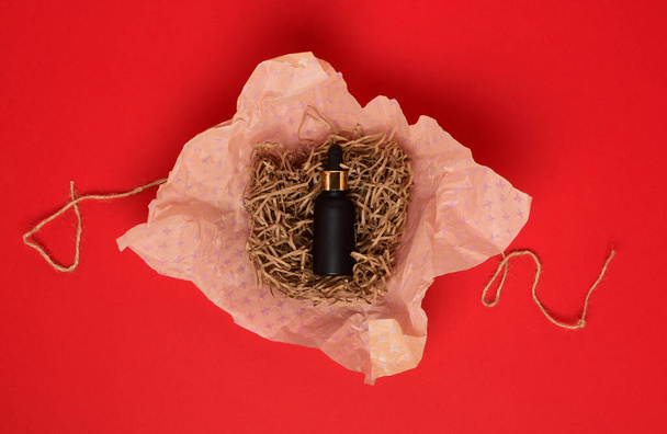 A bottle of black matte color with serum lies on a red background. The bottle lies on a beige festive wrapping paper. The concept of makeup products. Flat lay. - Valokuva, kuva