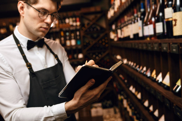 Professional cavist making notes at degustation card standing near wine shelves - Photo, image