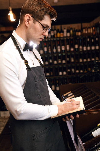 Wine steward looking at bottles of wine and writing in notebook at wine cellar - Zdjęcie, obraz