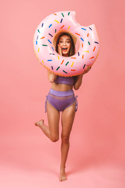 Full length portrait of a happy young woman dressed in swimsuit jumping and holding swim inflatable ring isolated over pink background. - Foto, Imagen
