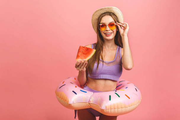 Portrait of a happy young woman dressed in swimsuit jumping and holding swim inflatable ring isolated over pink background. Holding watermelon. - Photo, Image