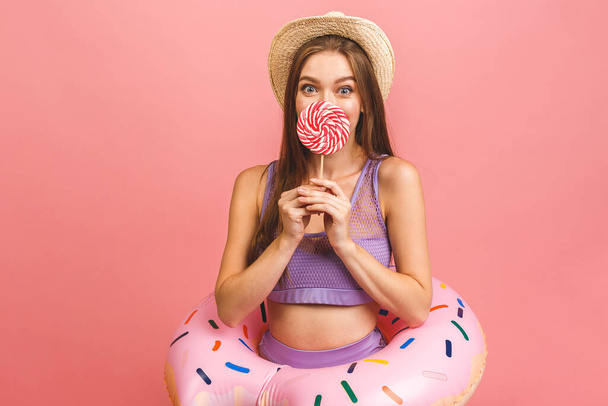 Porträt einer glücklichen jungen Frau im Badeanzug, die einen aufblasbaren Schwimmring isoliert vor rosa Hintergrund hält. Süßigkeiten in der Hand. - Foto, Bild