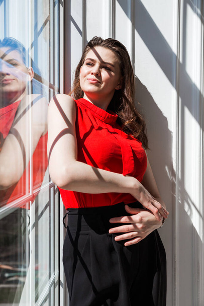 Menina morena na moda em uma blusa vermelha posa perto da janela e desfruta da luz solar e calor. Foto vertical - Foto, Imagem