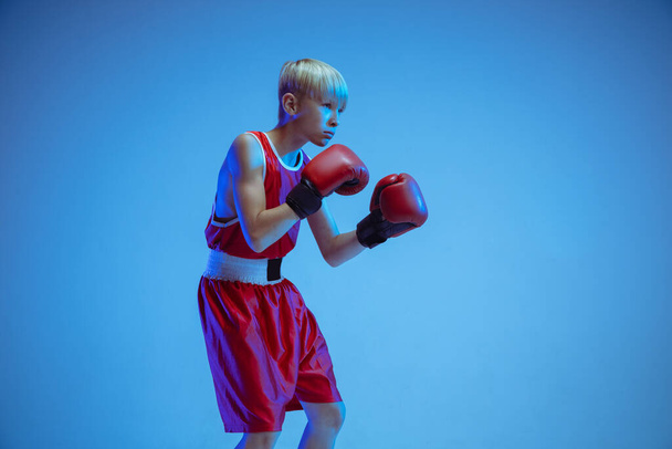 Teenager in sportswear boxing isolated on blue studio background in neon light - Foto, Imagem