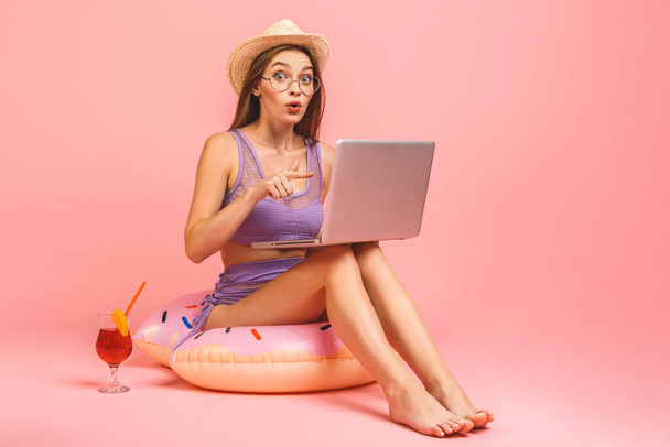 Vicces fiatal nő fürdőruhában, rózsaszín háttérrel. Az emberek nyaralás pihenés koncepció. Ülj úszás felfújható gyűrű, munka laptop PC számítógép. - Fotó, kép