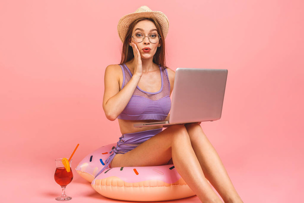 Funny young woman in swimsuit isolated on pink background. People summer vacation rest concept. Sit in swim inflatable ring, work on laptop pc computer. - Photo, Image