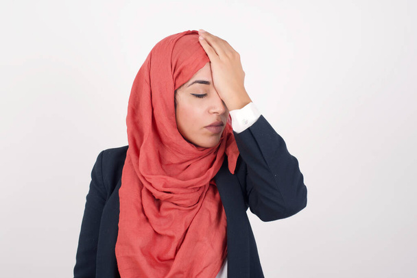 Frustrated Beautiful muslim woman  wears black blazer and hijab wearing casual clothes holding hand on forehead being depressed regretting what she's done having headache. Confused female with frowned face looking stressful having some pain  - 写真・画像