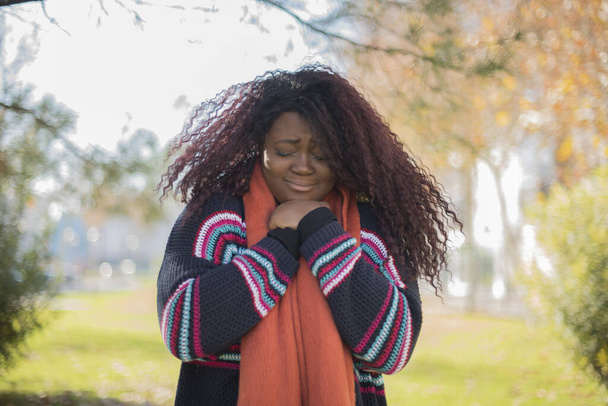カラフルなセーターを身に着けている長い髪の美しいアフリカ系アメリカ人女性は秋の公園で悲しいです   - 写真・画像