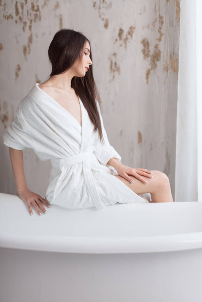 Beautiful young lady in bath robe going to take a bath in white bathroom - Photo, Image