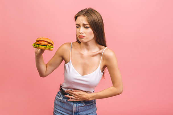 Die junge Frau leidet unter Bauchschmerzen. Sie hält einen gebissenen Burger in der Hand. Eine andere Hand liegt auf der Bauchgegend. Vereinzelt auf rosa Hintergrund. - Foto, Bild