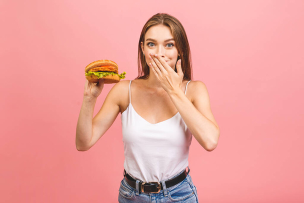 Πορτρέτο της νεαρής όμορφης πεινασμένης γυναίκας τρώει burger. Απομονωμένο πορτραίτο μαθητή με γρήγορο φαγητό πάνω από ροζ φόντο. Έννοια διατροφής.  - Φωτογραφία, εικόνα