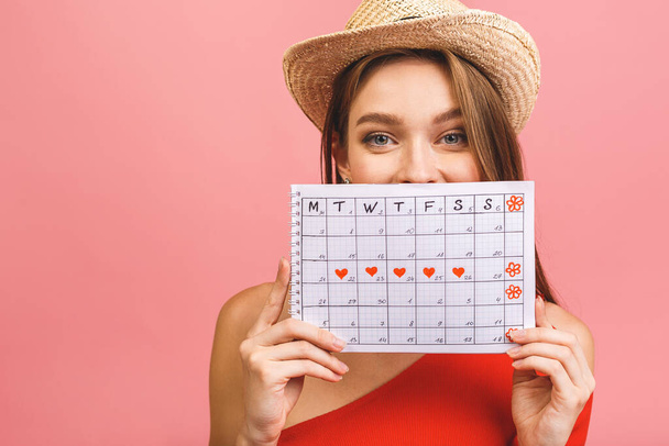 Portrait of a funny young girl in summer hat hiding behind a periods calendar isolated over pink background. - Photo, Image