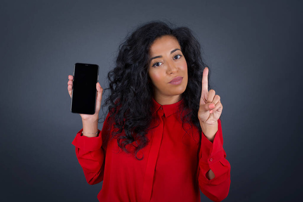 Young beautiful brunette woman with curly hair wearing red shirt with smartphone pointing up in gray studio  - Photo, image