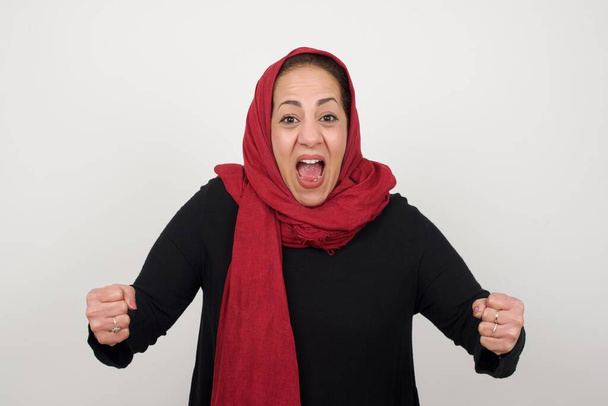 灰色の壁に立ってヒジャブを身に着けている成熟したイスラム教徒の女性の肖像画はカメラで興奮して見ます,手を頭の上に上げて,予期しないことに気づきます - 写真・画像