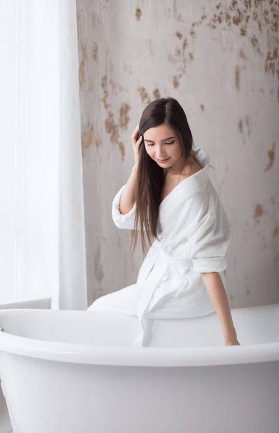 Spa european girl in white robe, preparing to bath, looking down at water - Fotografie, Obrázek